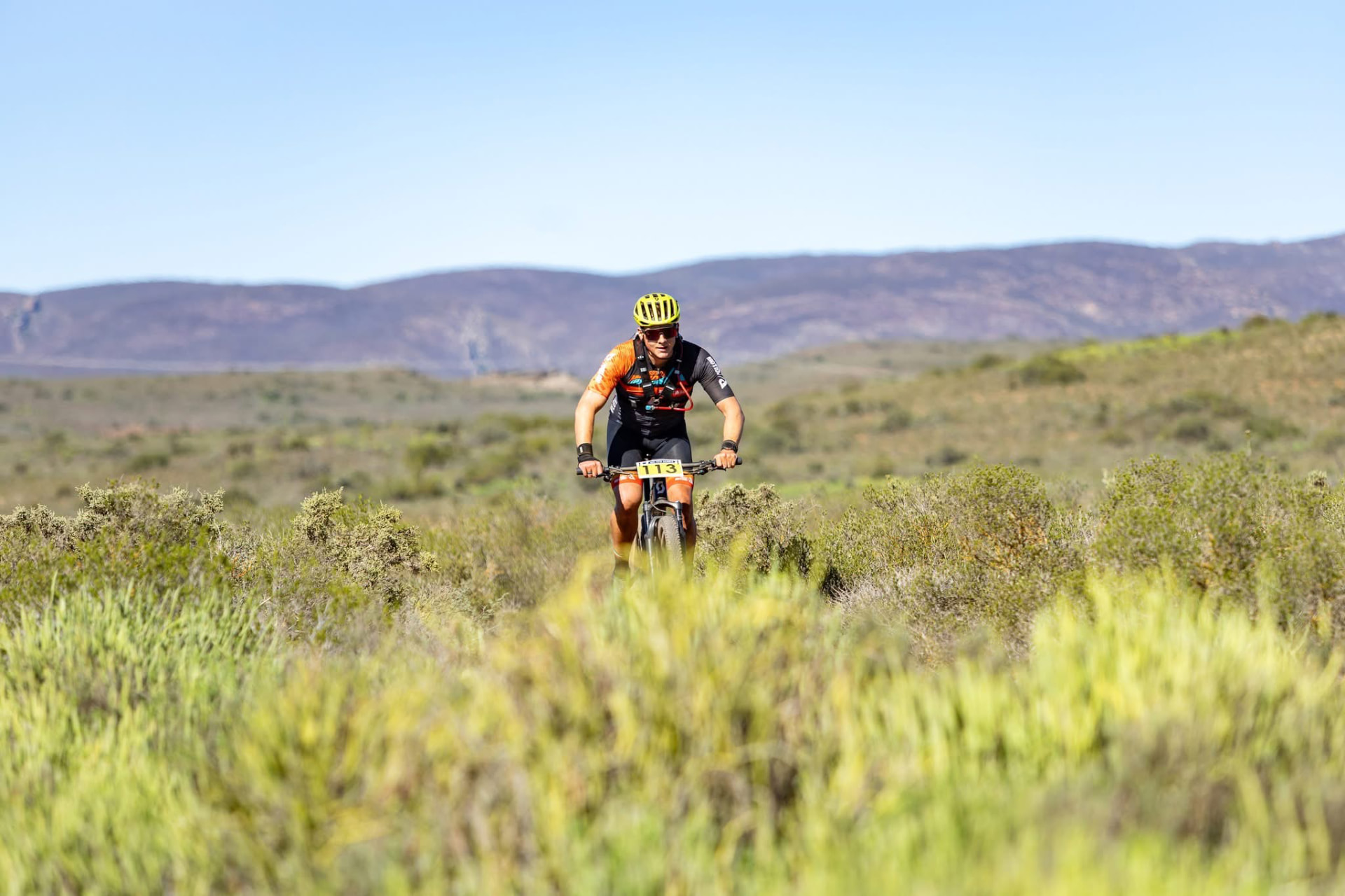 Dawid Oosthuizen racing marathon MTB through open Cape fynbos with mountains behind